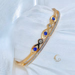 Gold bangle bracelet with navy Evil Eye and clear zirconia, dual-layer front design on soft white fabric background.