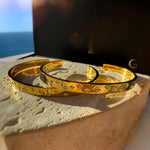 Two gold cuff bracelets with celestial star engravings and cubic zirconia, resting on a natural stone under warm light.