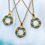 Three gold circle necklaces with heart details and blue crystals, arranged on soft fabric. Each pendant forms a wreath shape made of alternating hearts and sparkling blue stones.