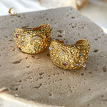 Close-up of large textured gold hoop earrings with moon, star, and evil eye symbols, placed on stone surface, soft natural light.