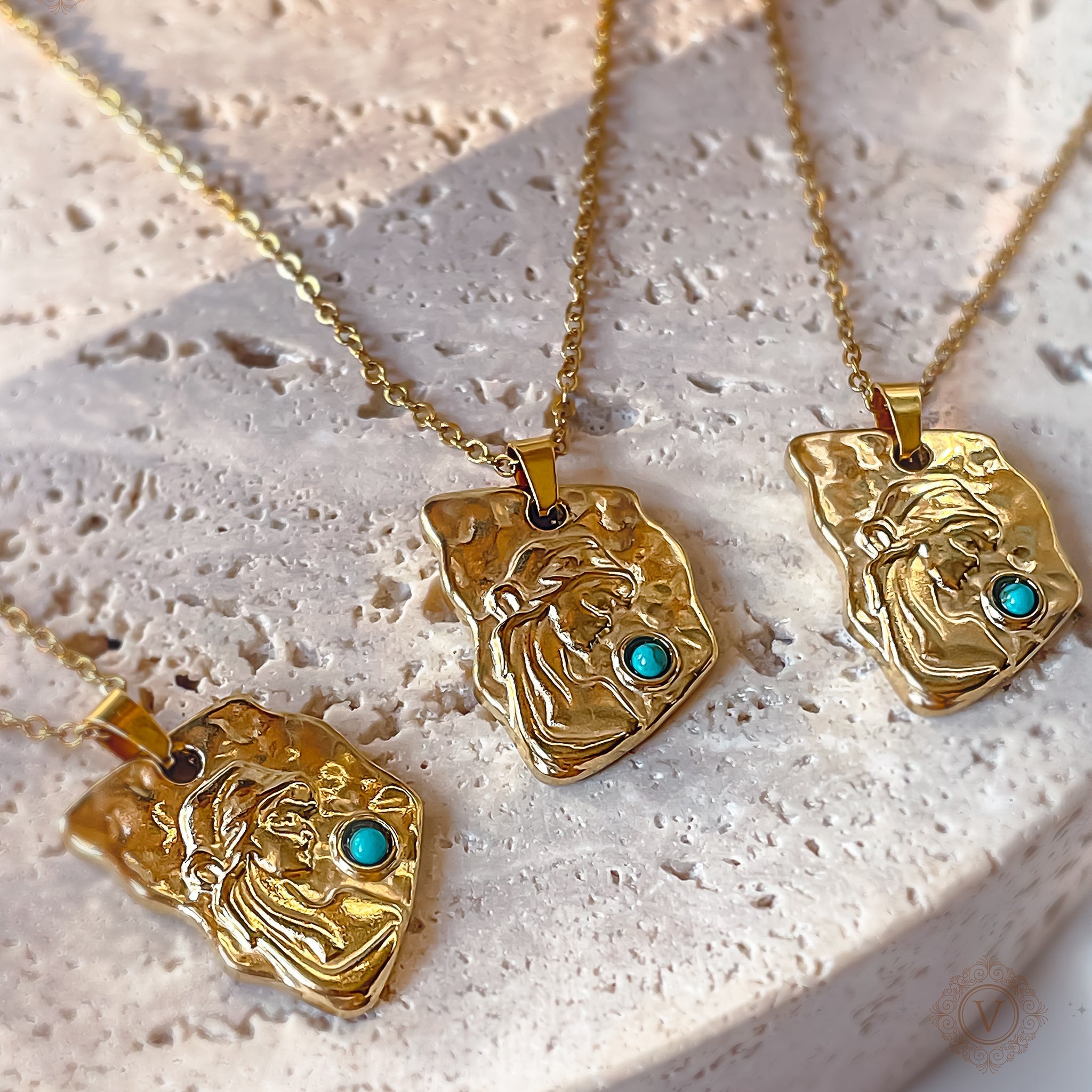 Three gold medallion necklaces featuring a woman’s face in a headscarf and a central turquoise stone, displayed on textured stone.