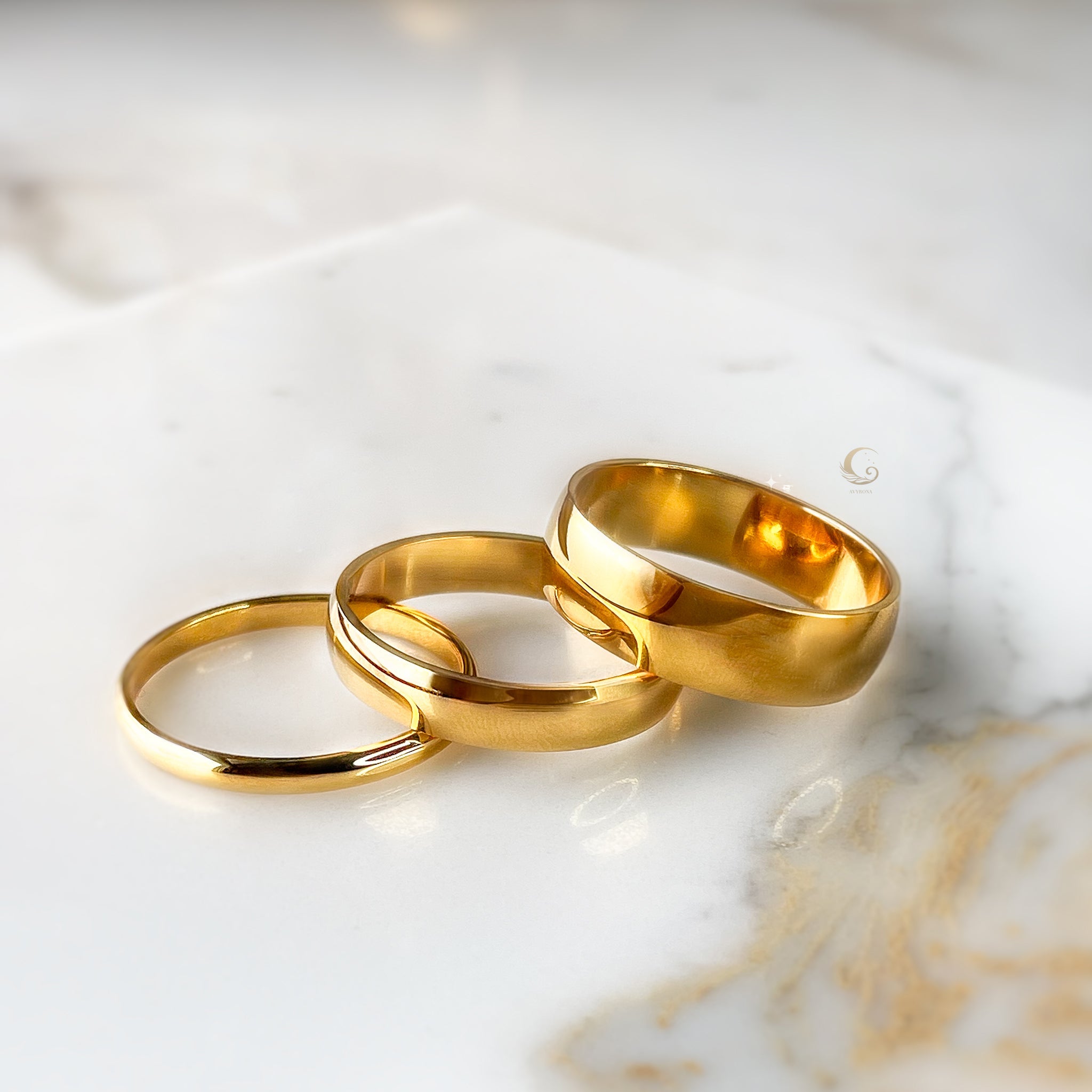 Three gold rings on a white marble background, representing classic and statement jewelry styles.