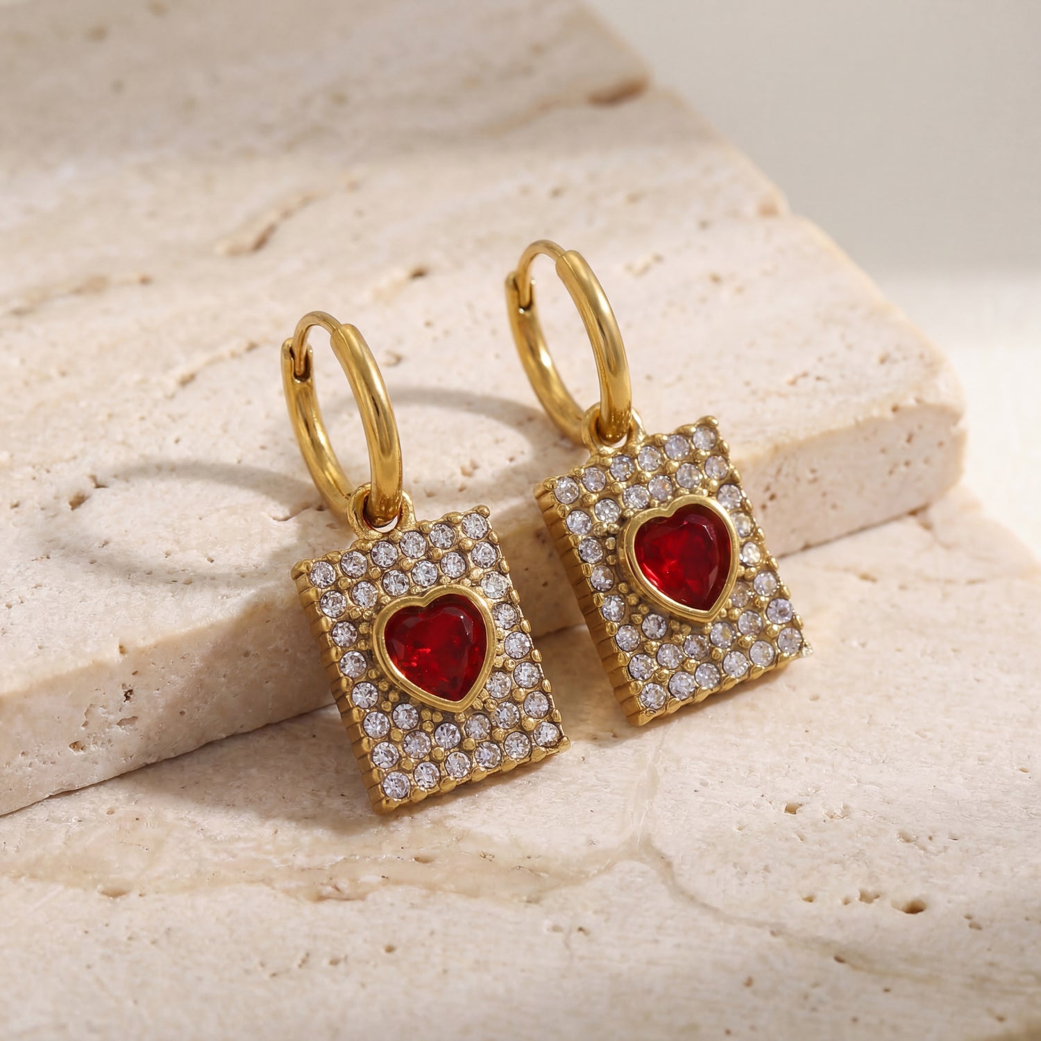 Gold earrings with red heart stones on a beige stone surface