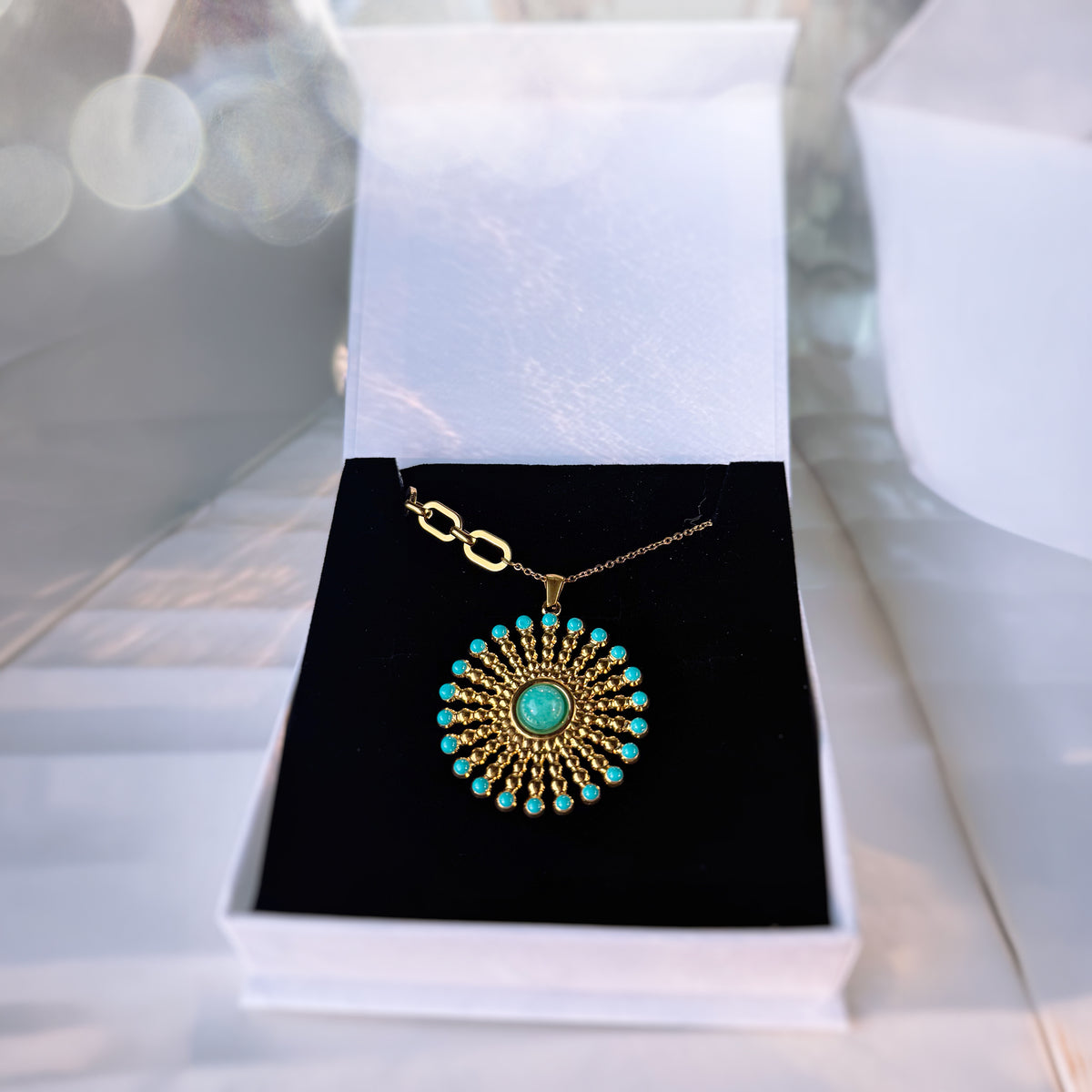 A gold pendant necklace with a turquoise center and surrounding turquoise stones, displayed on a black velvet insert inside an open white jewelry box.