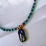 A beaded necklace with faceted green and black beads, featuring a gold-toned pendant with a crescent moon and raised hand design on a dark blue-green background.