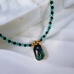 A beaded necklace with green and black faceted beads and a gold-toned pendant featuring a teal and green religious icon, displayed on a white marble surface.