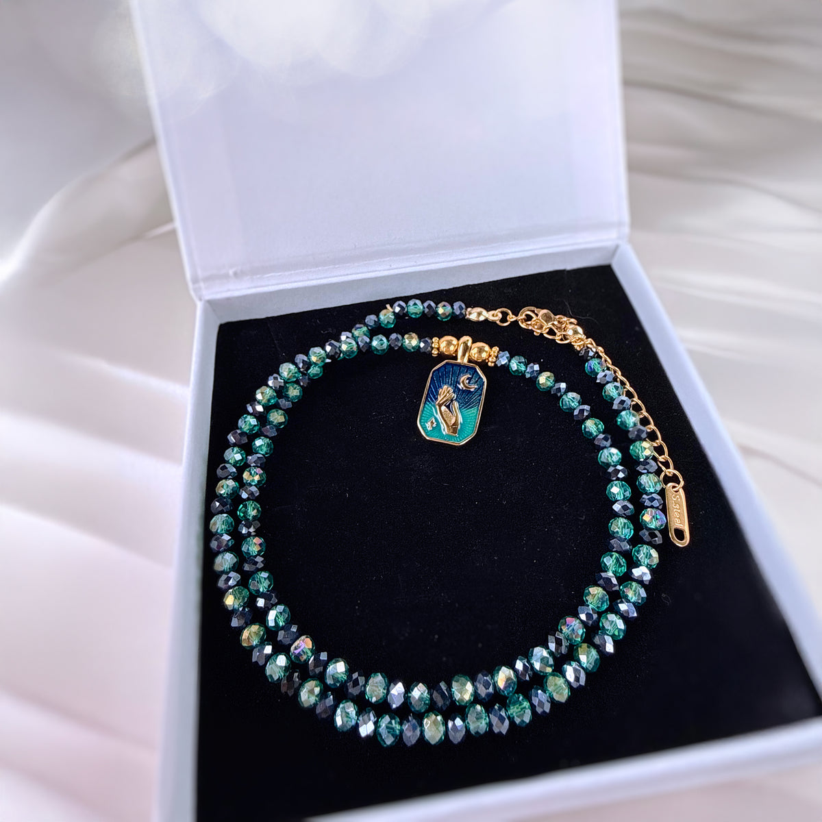 A beaded necklace with iridescent green and blue faceted beads, gold-tone chain links, and a small rectangular enamel charm, displayed inside an open white jewelry box with a black velvet insert.