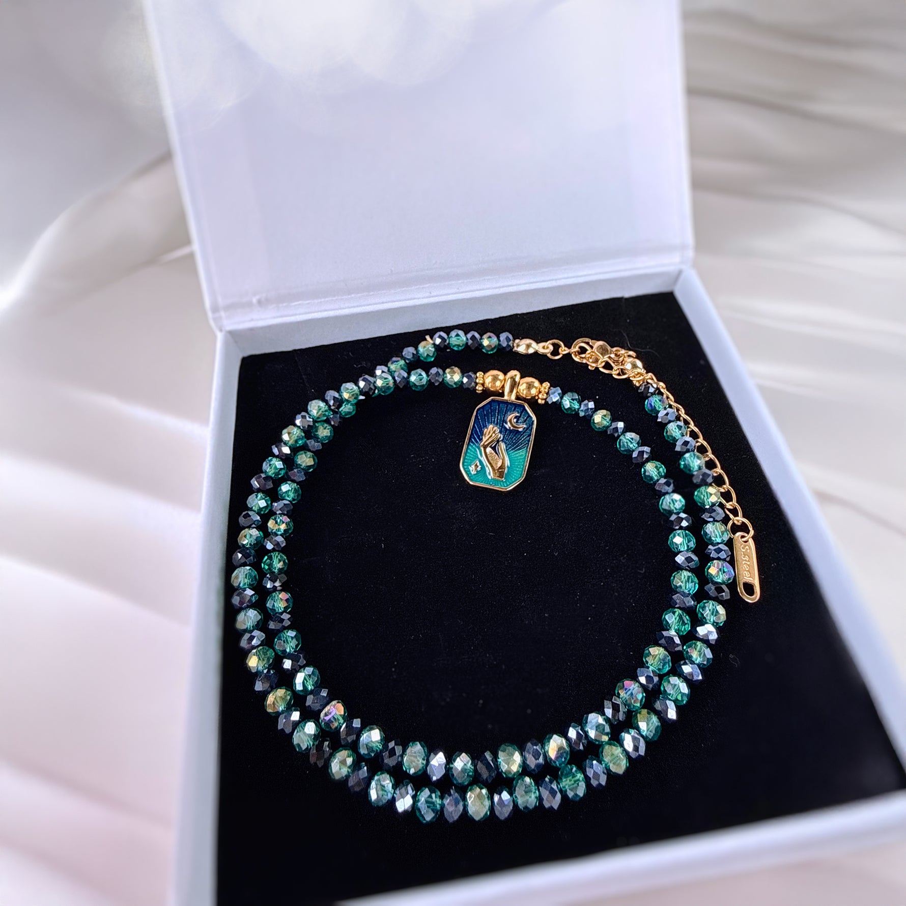 A beaded necklace with iridescent green and blue faceted beads, gold-tone chain links, and a small rectangular enamel charm, displayed inside an open white jewelry box with a black velvet insert.