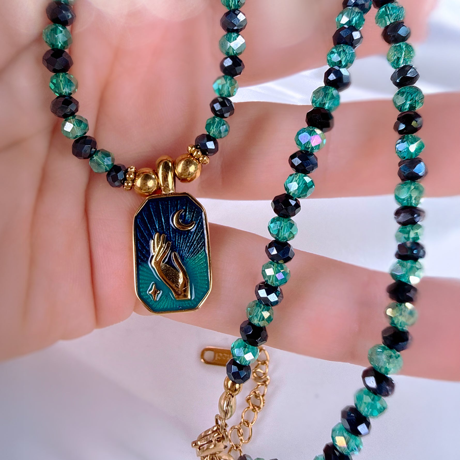 A close-up of a beaded necklace draped over a hand, featuring faceted green and black beads with a gold-tone pendant. The pendant has a dark blue and teal background with a gold crescent moon and raised hand design.