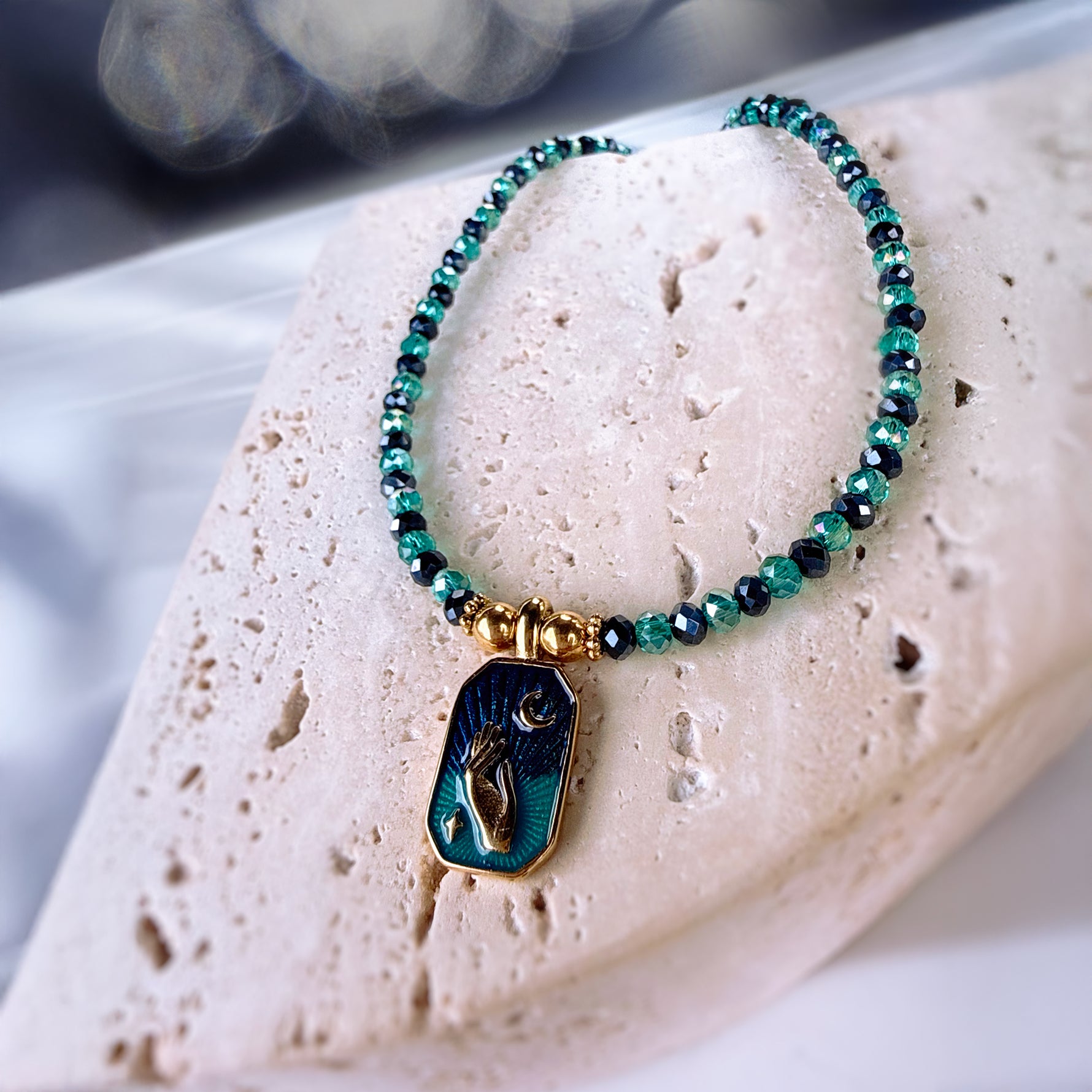 A beaded bracelet with green and black faceted beads, gold-toned spacers, and a rectangular pendant featuring a gold-toned figure on a dark blue and green background, displayed on a light stone surface.