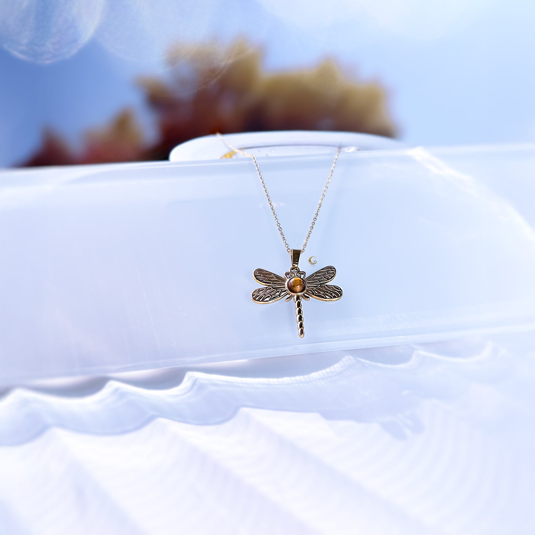 Gold dragonfly necklace on a white stand with a blurred floral background