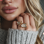 A close-up lifestyle shot of a woman wearing a gold ring with three flower-shaped elements, one of which is open to reveal a small white gemstone. She is wearing a gray knit sweater.