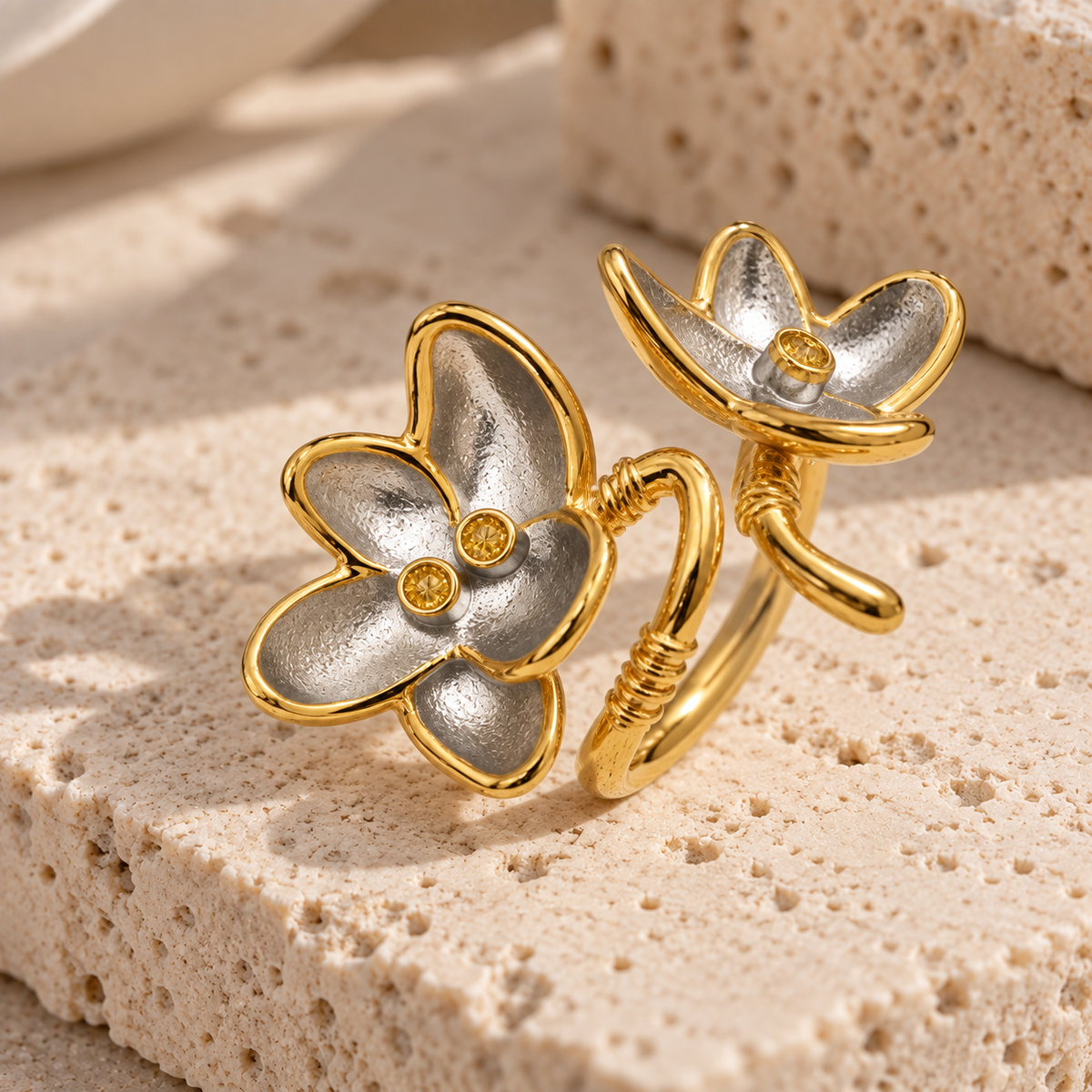 A pair of gold and silver flower-shaped rings displayed on a textured beige stone surface.
