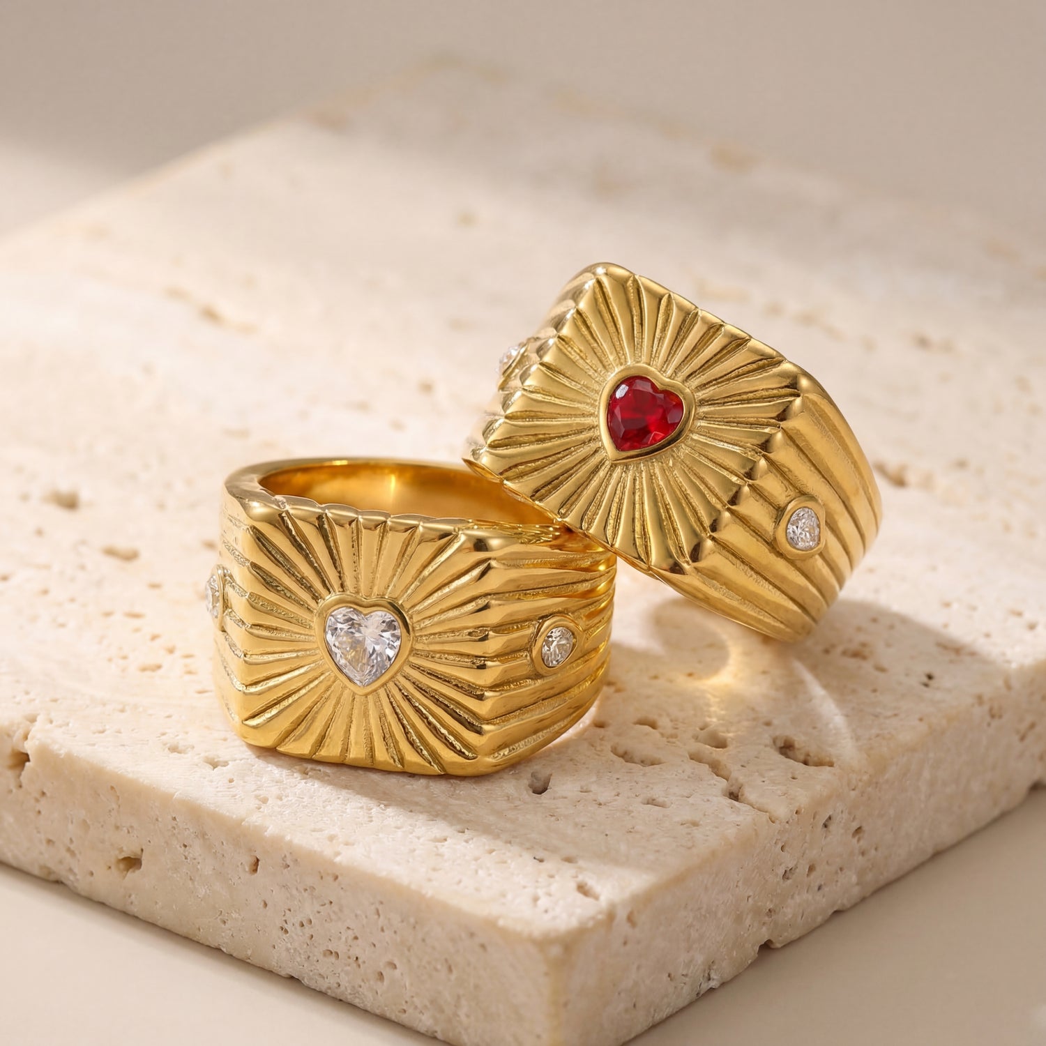 Two gold rings with heart-shaped gemstones on a beige stone surface