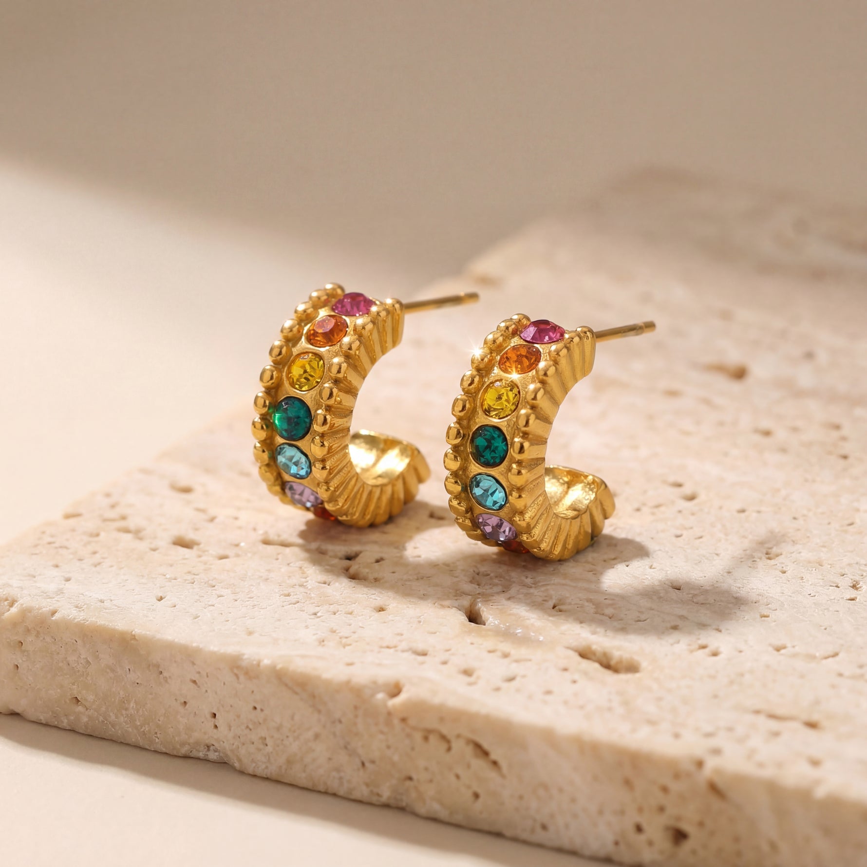Gold hoop earrings with colorful gemstones on a beige stone surface