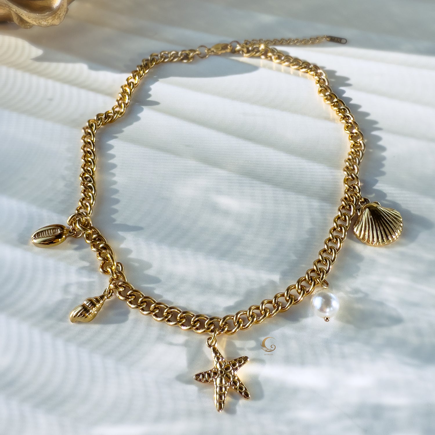 Golden charm necklace with shells, vegan pearl and star arranged on textured stone plate with wave-like pattern