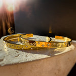 Golden starry cuff bracelets placed on textured beige stone, glowing in a dreamy sunset ambiance.