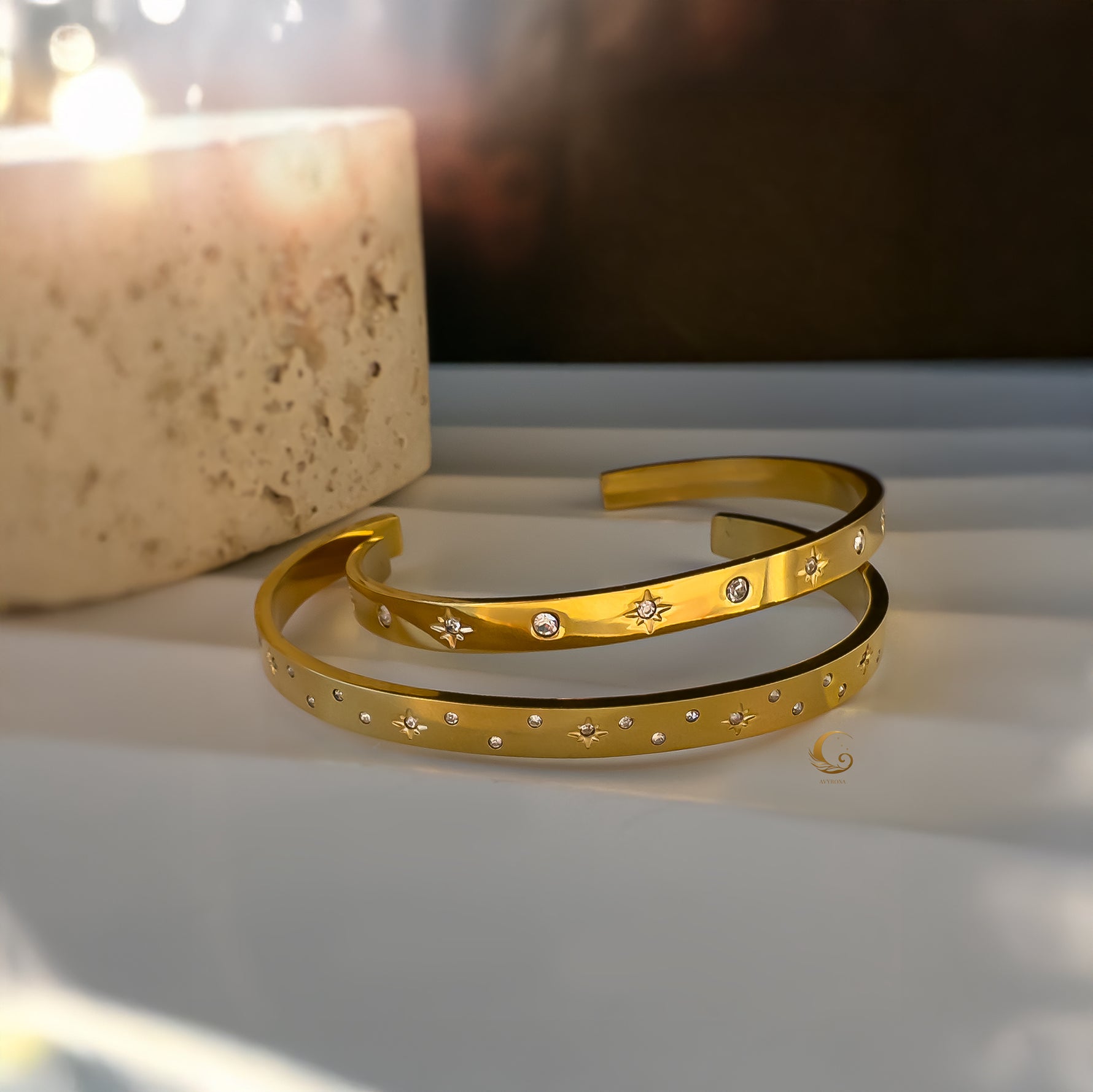 Two golden starry bracelets next to a candle, displayed on a soft neutral surface in warm light.