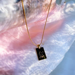 Gold necklace with a black pendant featuring a moon and hand design on a textured pink surface.