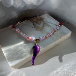 A beaded necklace with a glossy purple teardrop pendant, displayed on a white marble block. The necklace features light pink and white marbled beads, small gold spacer beads, and red accent beads.