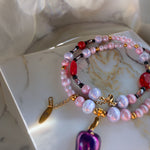 A close-up of a beaded bracelet or layered jewelry piece resting on a white marble tray. The jewelry features iridescent light pink and lavender beads, red faceted beads, small gold spacer beads, and gold-toned chain hardware with a purple pendant charm.