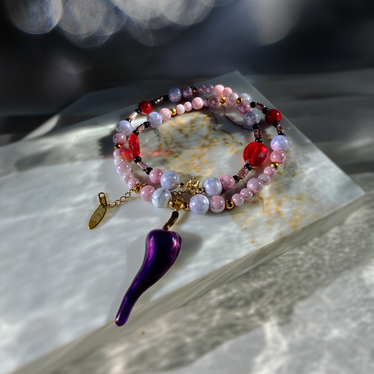 A beaded bracelet with pink, purple, and red beads, gold-toned chain links, and a dangling metallic purple charm, displayed on a white marble slab with dramatic light and shadow.