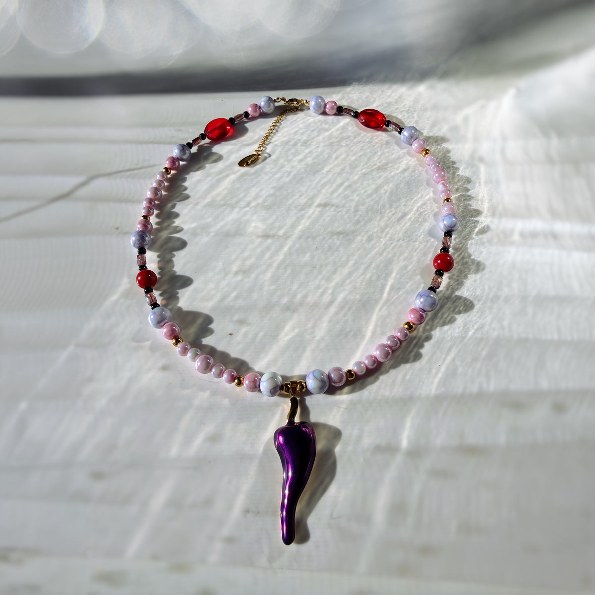 A beaded necklace with a glossy purple chili pepper pendant, featuring pink, red, and lavender beads with gold-toned metal accents, displayed on a textured white fabric background.