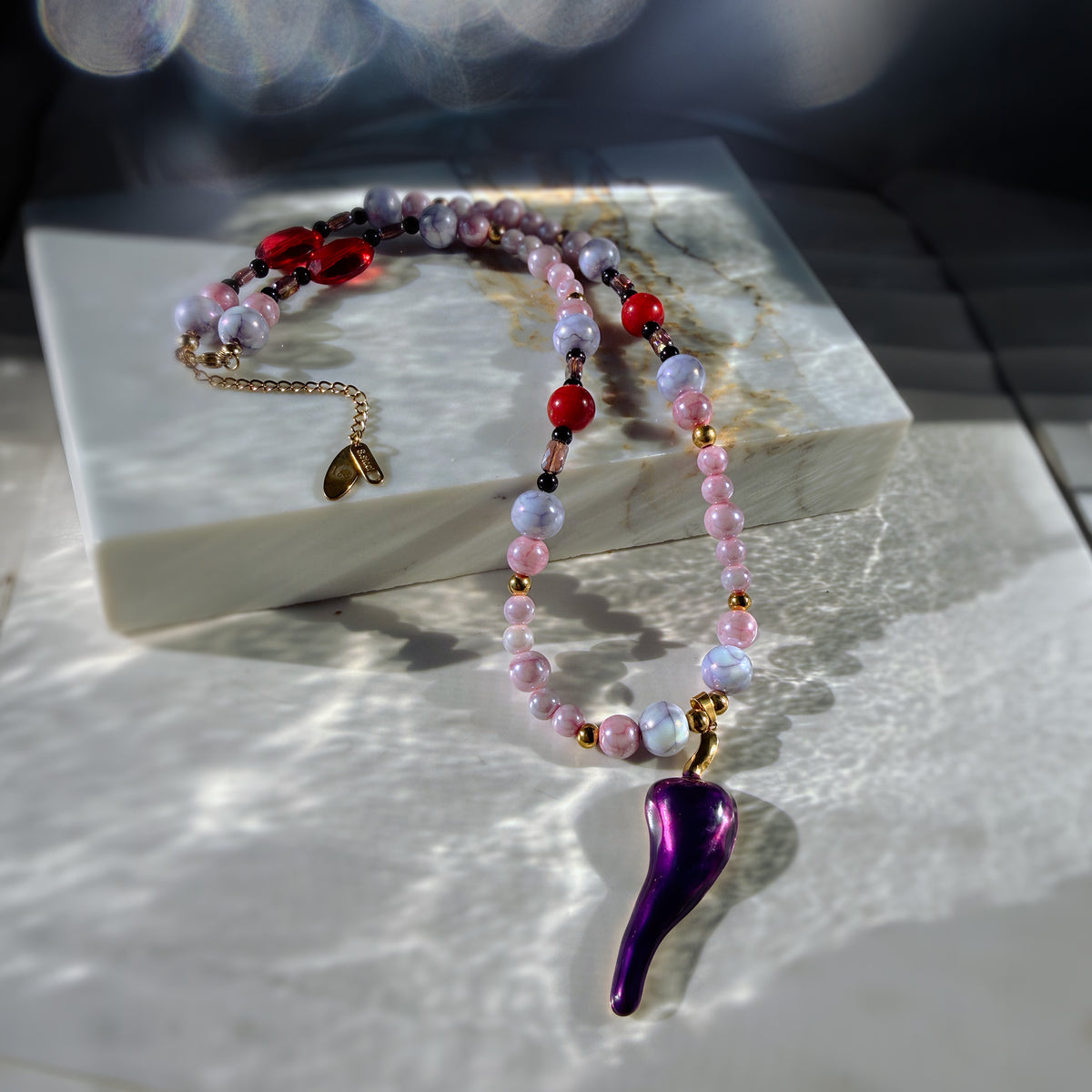 A beaded necklace with pink, lavender, and red beads, gold-toned spacers, and a glossy purple teardrop-shaped pendant, displayed on a white marble block with dramatic natural light shadows.