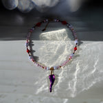 A beaded necklace with light pink and white beads, small red accents, and a glossy purple teardrop-shaped pendant, displayed on a white surface with dramatic sunlight and shadows.