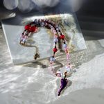 A beaded necklace with pink, purple, and red beads, gold-toned chain details, and a glossy purple chili pepper pendant, styled on a white marble block with dramatic natural light and bokeh.