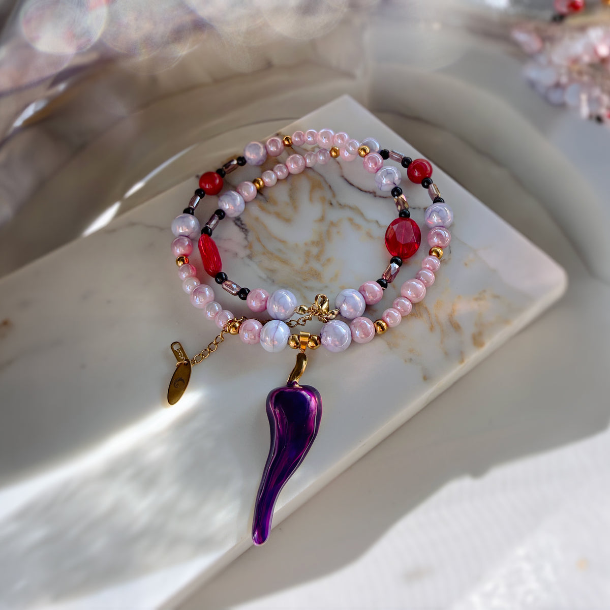 A beaded bracelet set styled on a white marble tray. The set features light pink and white marbled pearls, red faceted beads, black spacer beads, gold-toned metal spacers and chain, and a prominent purple glass chili pepper charm.