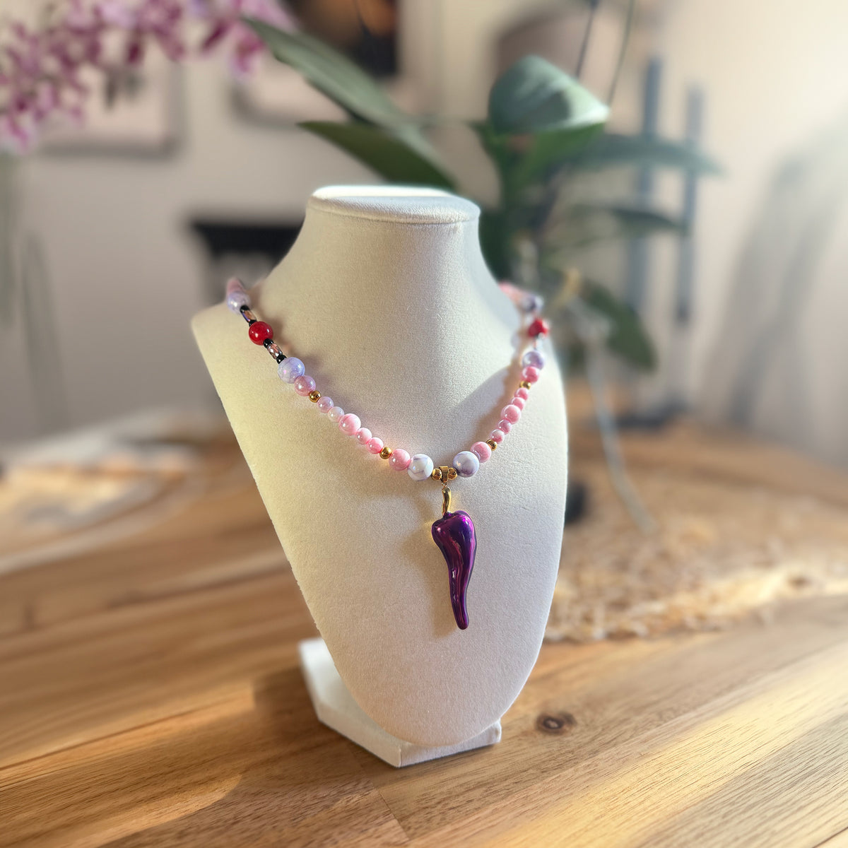 A beaded necklace with pink, white, and red beads and a purple teardrop-shaped pendant, displayed on a white jewelry bust on a wooden table.