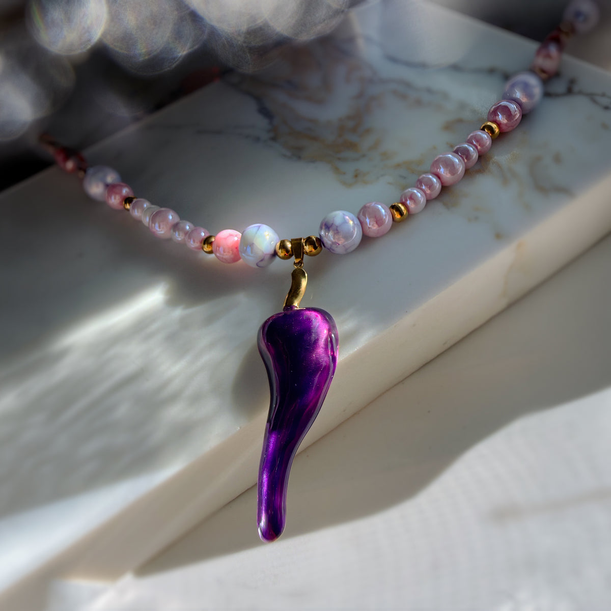 A beaded necklace with iridescent pink and white beads, small gold spacer beads, and a glossy metallic purple teardrop-shaped pendant, displayed on a white marble slab.
