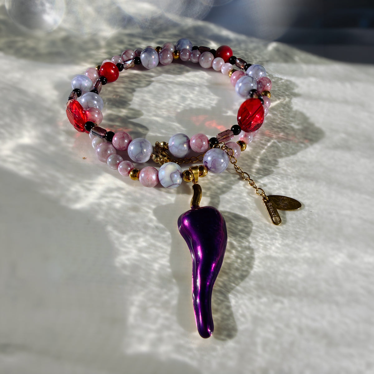 A beaded bracelet with red, pink, and lavender marbled beads, a gold chain, and a dangling metallic purple chili pepper charm, displayed on a textured white surface with dappled sunlight.