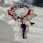 A beaded bracelet with red, pink, and lavender marbled beads, a gold chain, and a dangling metallic purple chili pepper charm, displayed on a textured white surface with dappled sunlight.