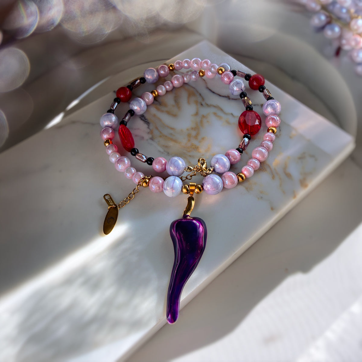 A beaded bracelet with iridescent pink and white beads, red faceted beads, gold-toned spacers, and a large purple teardrop pendant, displayed on a white marble slab.