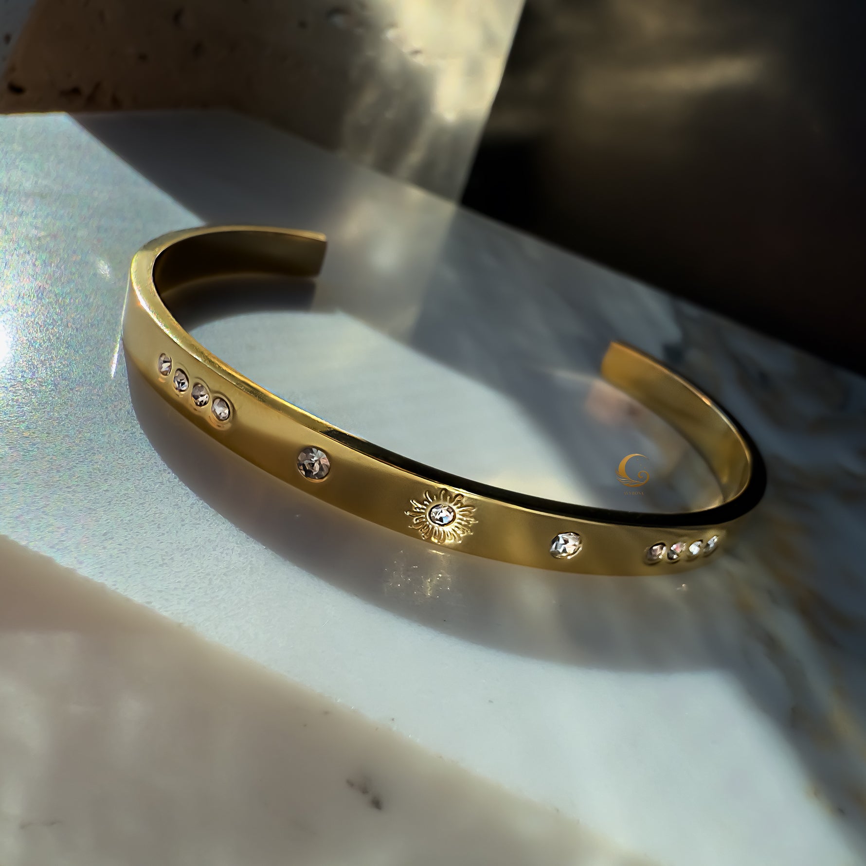 A delicate gold-plated bracelet with a minimalist sun charm and sparkling cubic zirconia stones, set against a wavy white stone background.