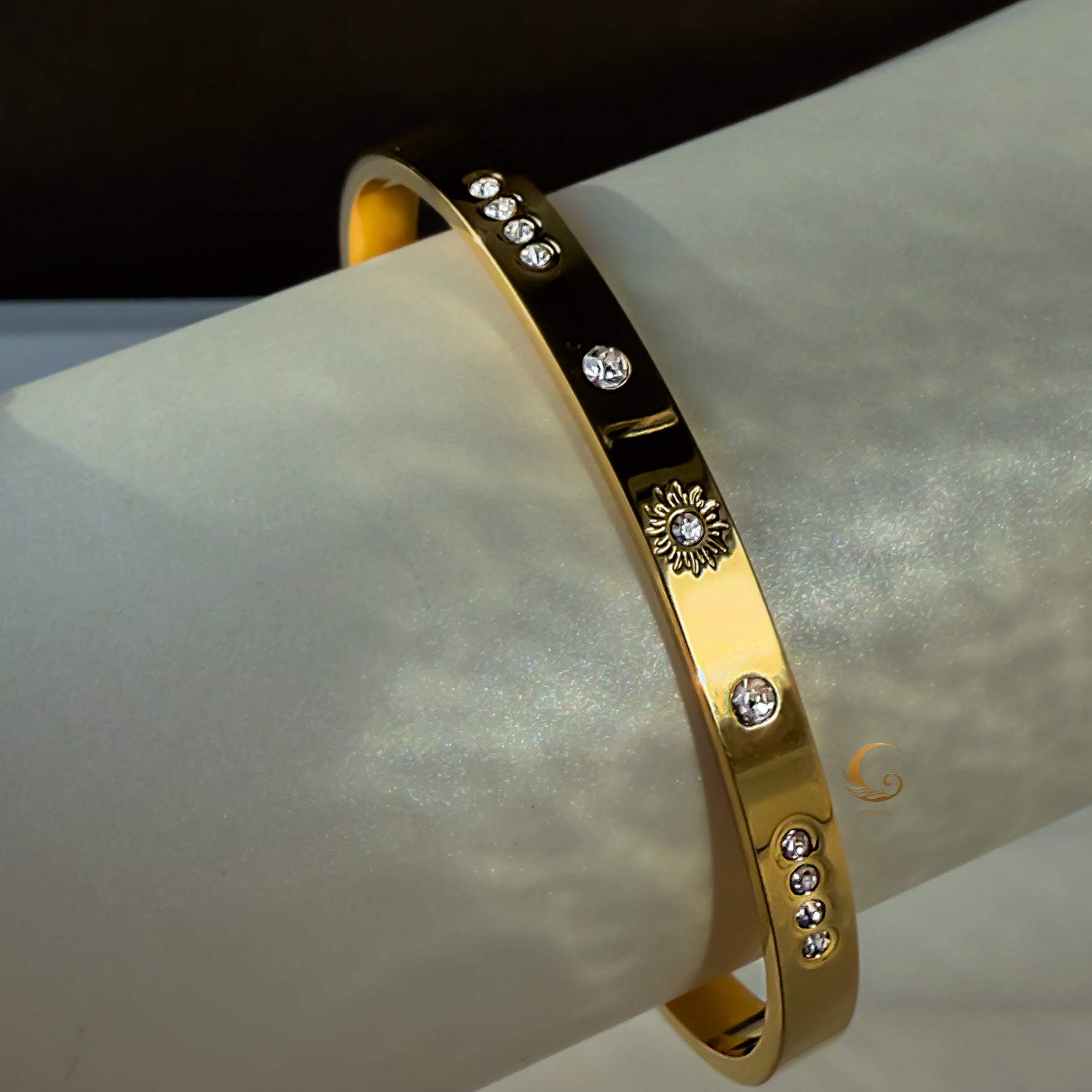 A gold bracelet with a sun motif and small cubic zirconia stones, displayed on a smooth, light-colored tube.