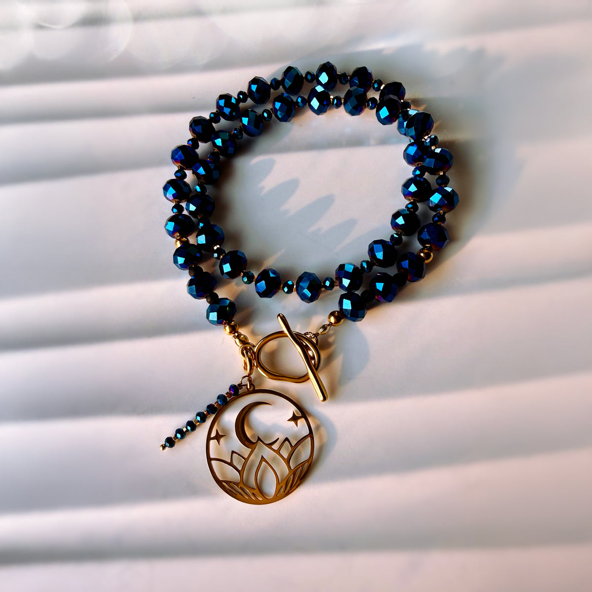 A beaded stretch bracelet with dark blue faceted beads and a gold-toned toggle clasp, featuring a dangling crescent moon and stars charm, displayed on a white slatted surface.