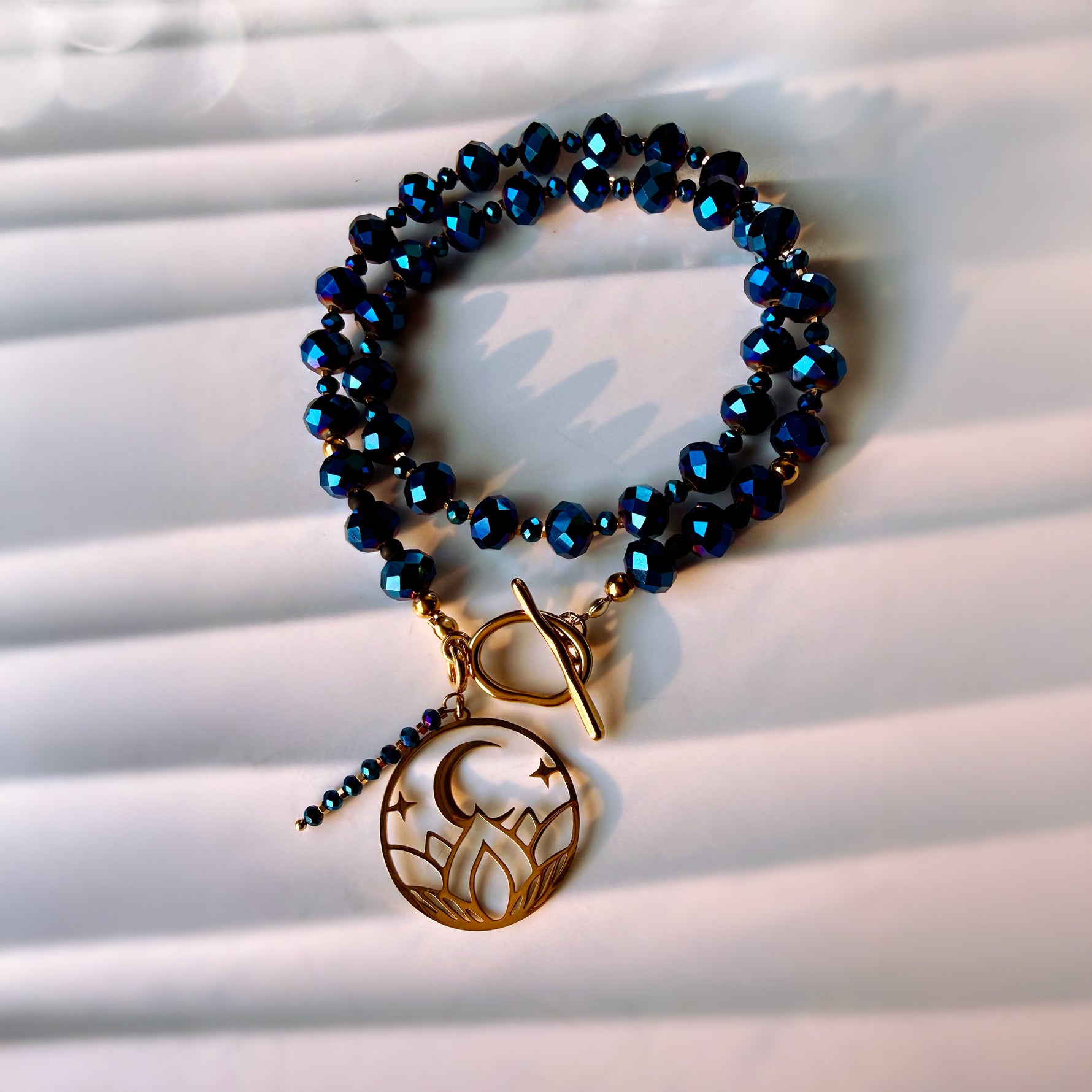 A beaded stretch bracelet with dark blue faceted beads and a gold-toned toggle clasp, featuring a dangling crescent moon and stars charm, displayed on a white slatted surface.