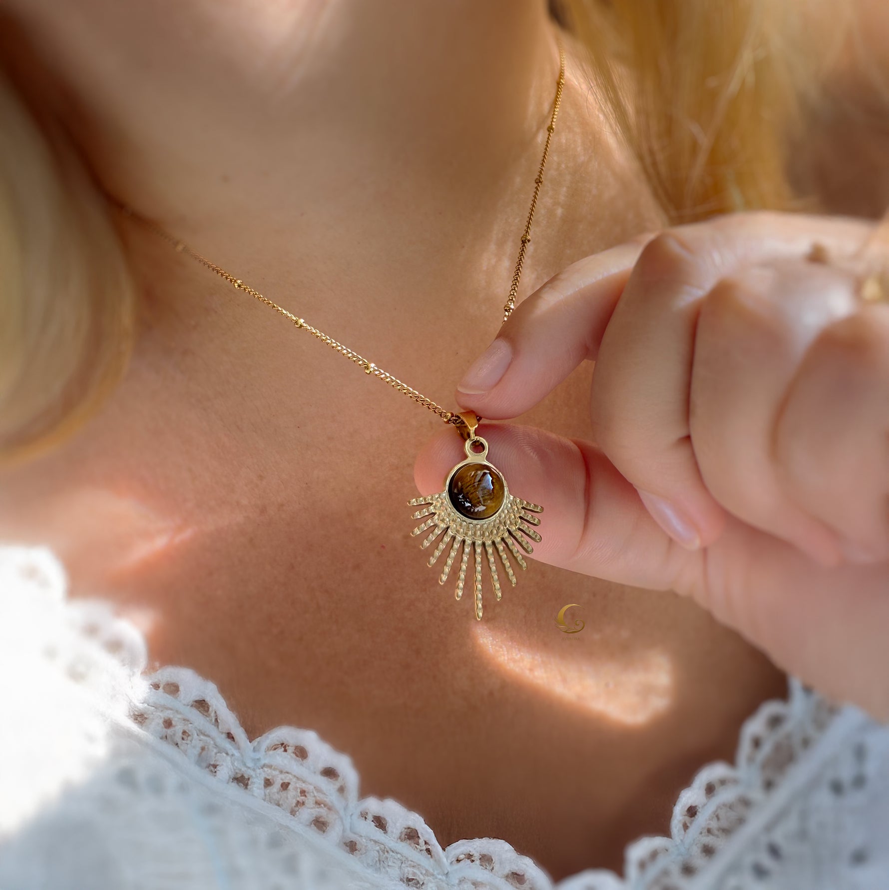 Close-up of a gold necklace with a sun pendant held by a person wearing a white lace garment.