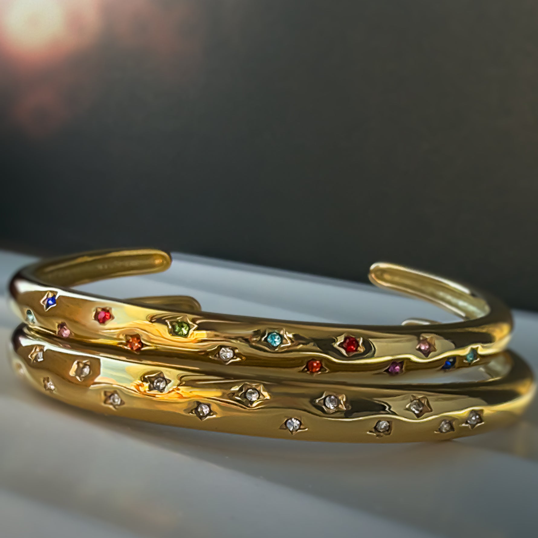 Close-up of Aerina gold cuff bracelets with rainbow and clear zircon star details, stacked together in soft light.