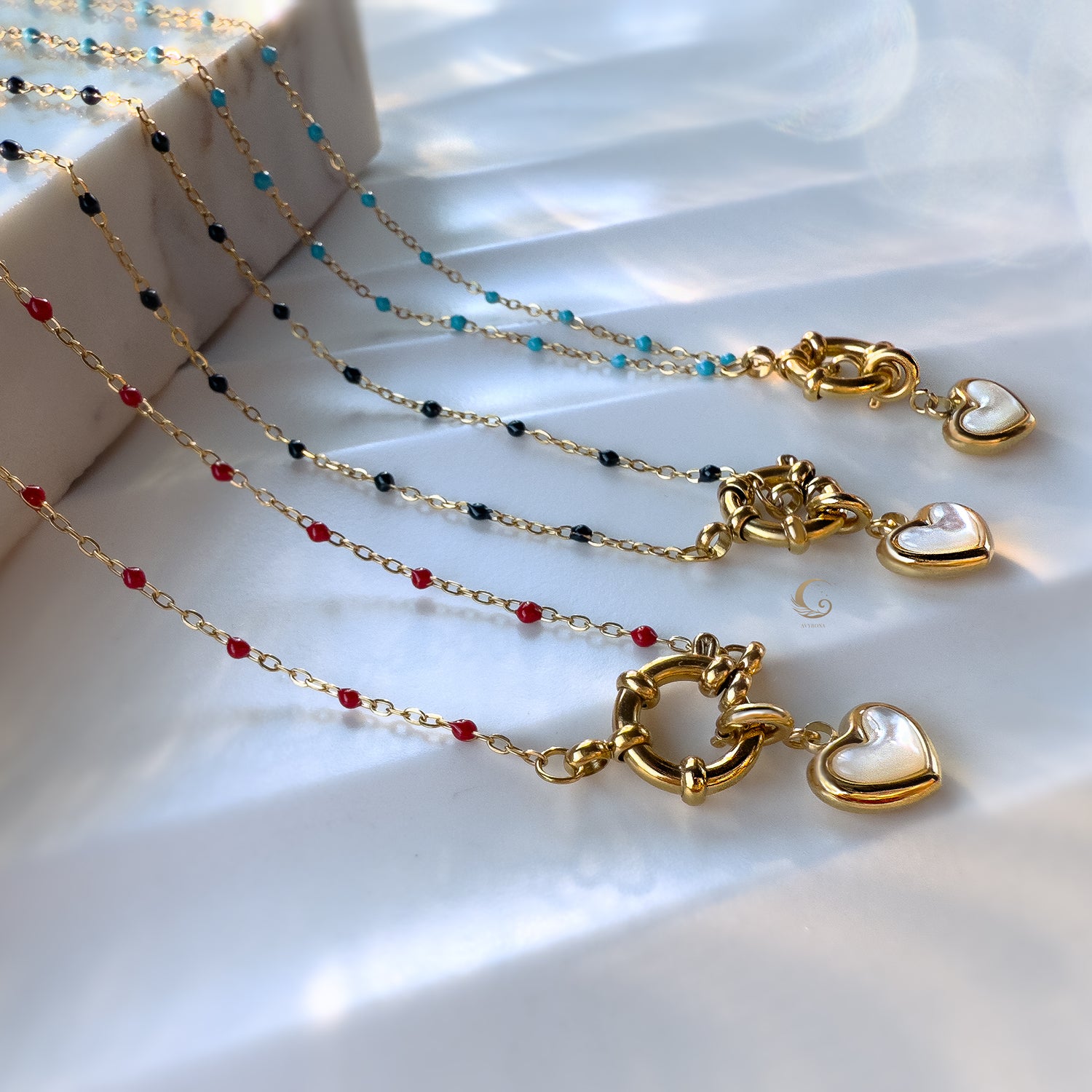  Four beaded necklaces in different colors, each with a gold toggle clasp and heart-shaped mother of pearl charm, arranged diagonally on a marble-effect surface with soft natural lighting.
