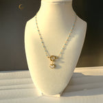The Alia necklace displayed on a white jewelry bust, highlighting its soft blue beaded chain and central gold heart charm made of mother of pearl, photographed in natural daylight.