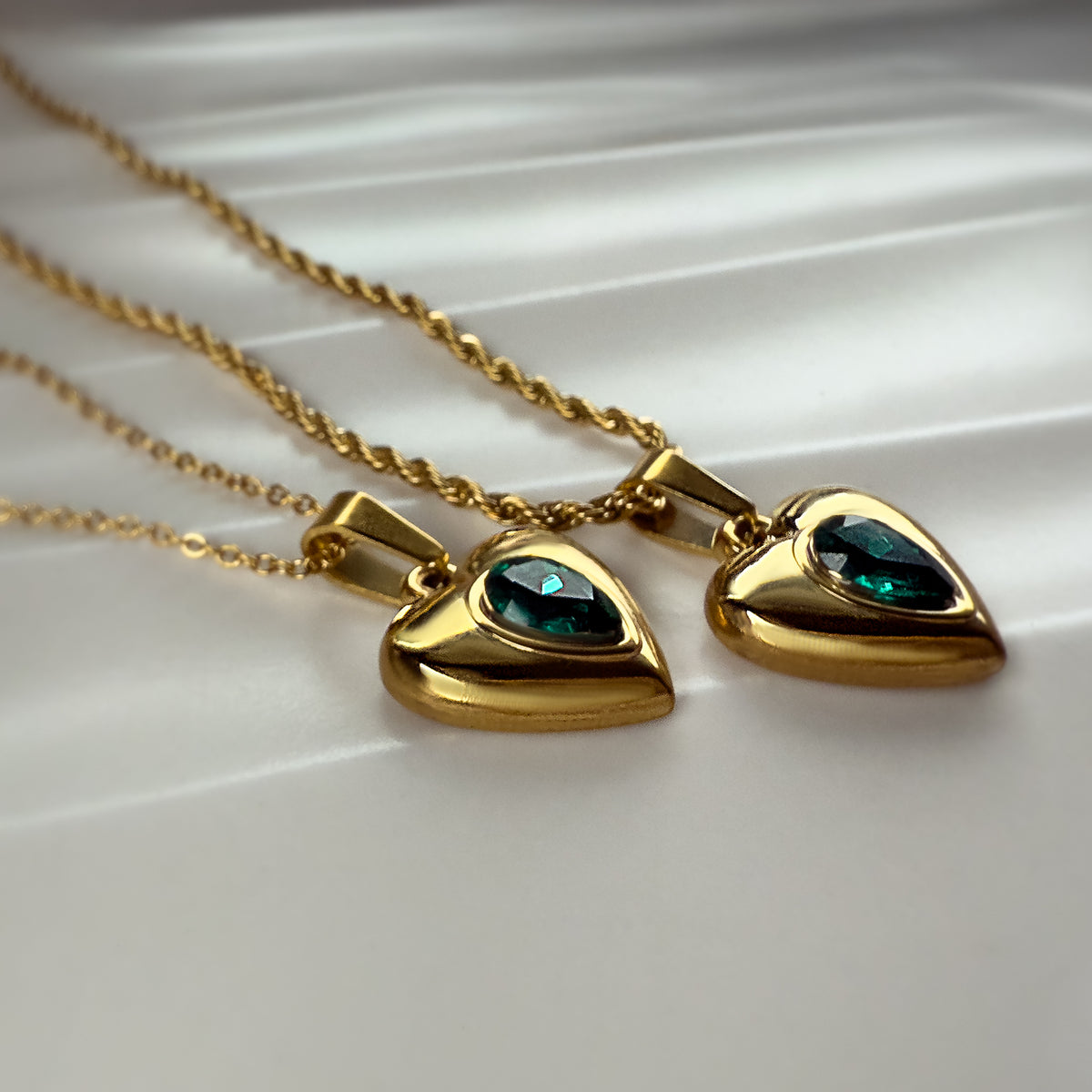  Emerald heart necklaces resting on white fabric, photographed at a side angle