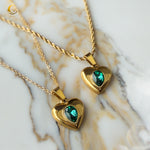 Close-up of two emerald heart necklaces with gold chains resting on beige marble, showing link and twisted chain variants