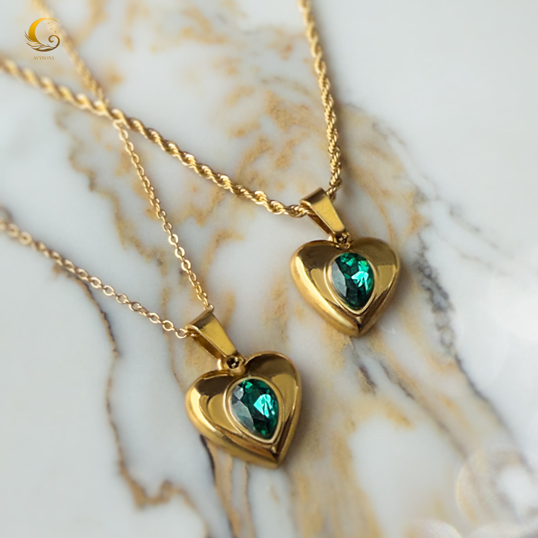 Close-up of two emerald heart necklaces with gold chains resting on beige marble, showing link and twisted chain variants