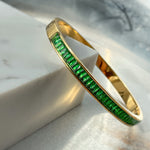 Emerald green crystal bangle bracelet in gold finish, displayed on white marble block – bold and radiant Fire Collection design by Avyrona
