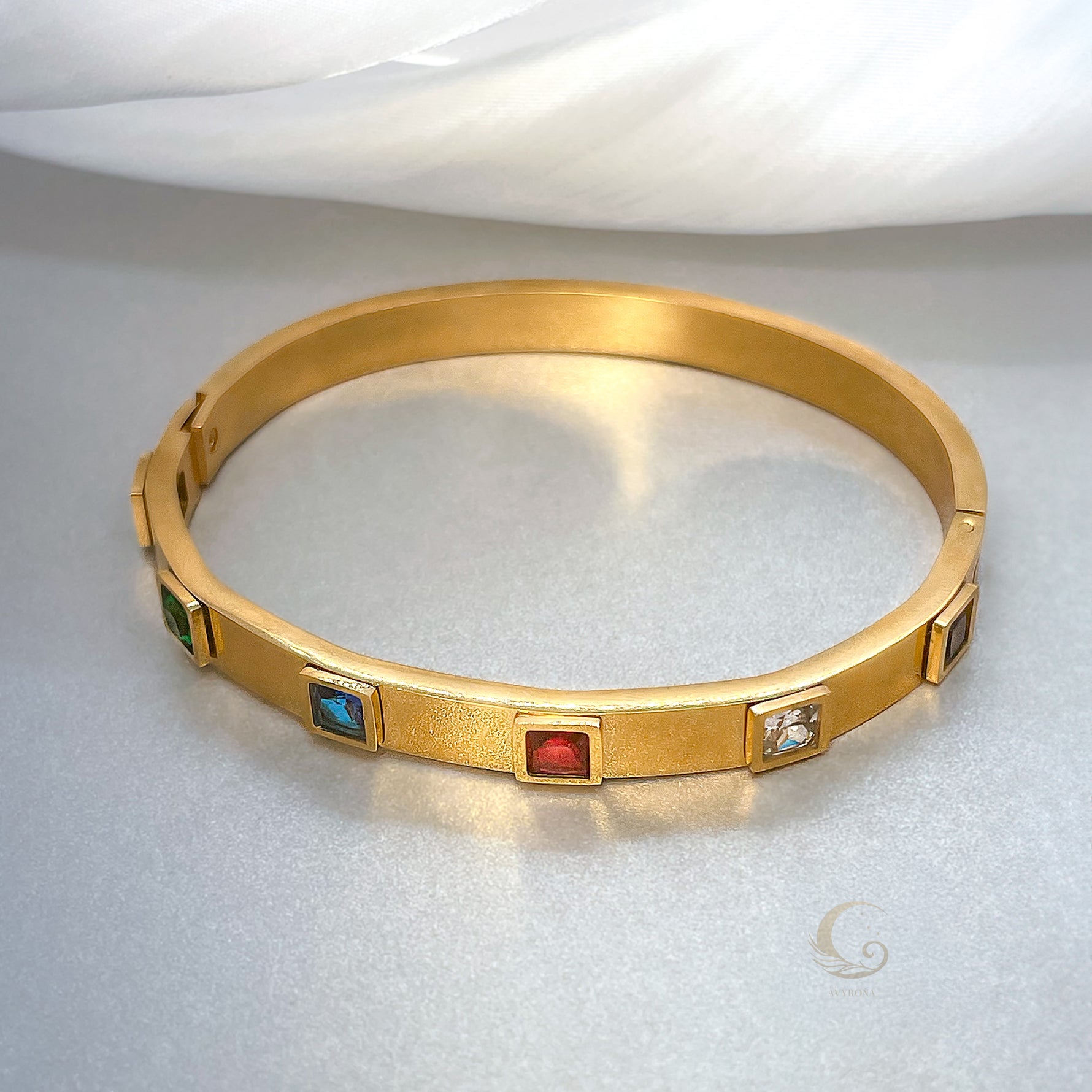 Full view of gold bangle bracelet with red, blue, green, and clear crystals, side clasp detail, placed on soft satin – Avyrona Fire Collection