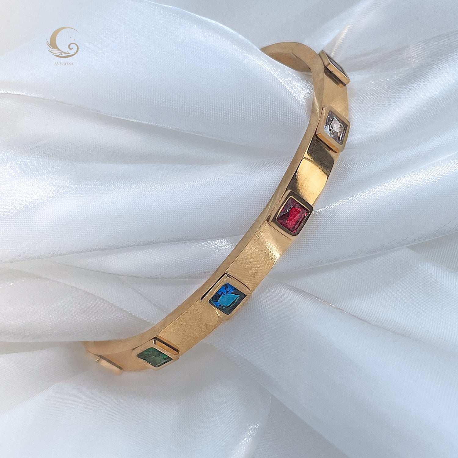 Elegant gold bracelet on satin background with colorful square crystals, featuring emerald green, sapphire blue, ruby red, and clear stones – Avyrona